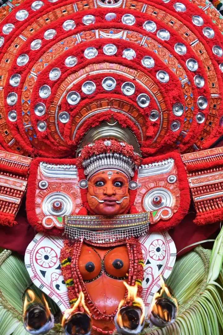 A Theyyam performer in a red headdress holding torches